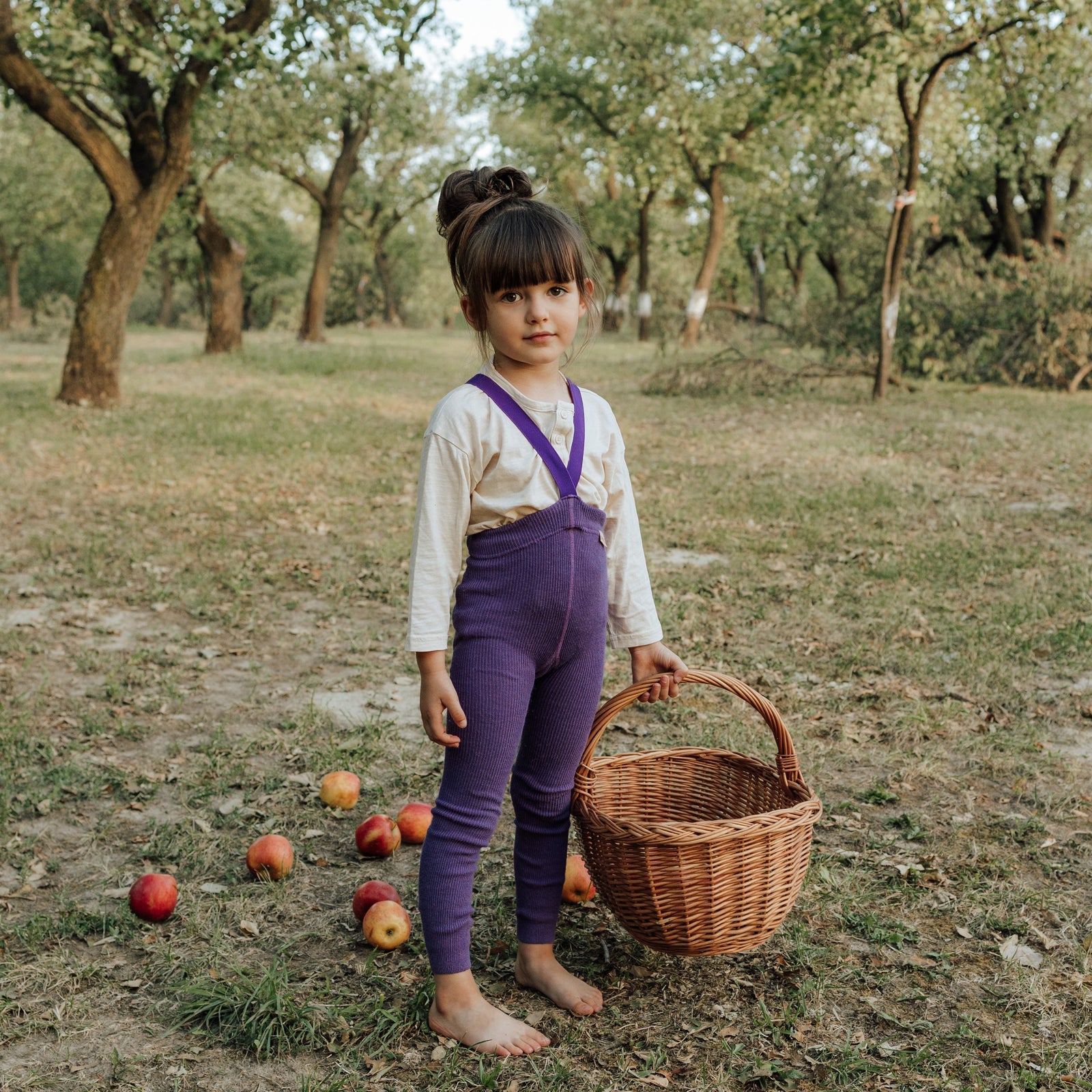 A little girl outside, wearing purple Peqne tights, holding a basket in her hand near some fallen apples.