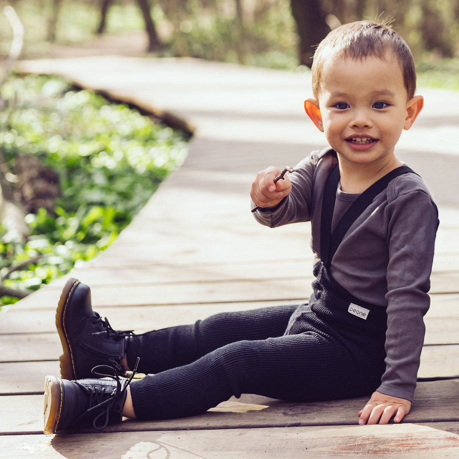 A photo of a small child outdoors, sitting on a wooden platform, wearing black Peqne tights with suspenders and shoes, pointing at the camera.